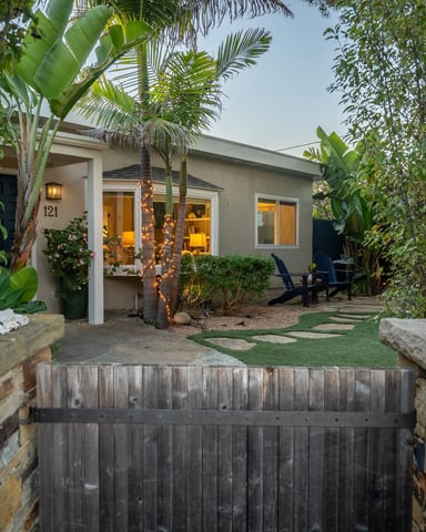 Garden-framed coastal cottage at dusk with fairy lights on a palm tree and a stone gate entrance