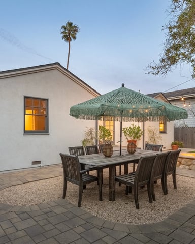 Backyard dining table under a macramé umbrella at dusk, with a stone fire pit and string lights