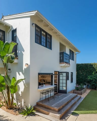 White two-story home with black-framed windows, banana leaf plants, and an outdoor kitchen bar in the garden