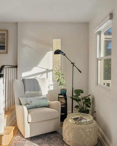 Sunlit reading nook with a white armchair, floor lamp, bookshelf, and windows overlooking the garden